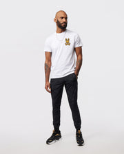 A man stands against a plain background, wearing a Psycho Bunny MENS VEGAS CITY TEE - B6U967X1PC made of Pima cotton jersey with a small printed bunny logo on the chest. He pairs it with black pants and black sneakers featuring golden accents. Tattoos adorn his arms, and he sports a beard and a short haircut, looking slightly to the side.