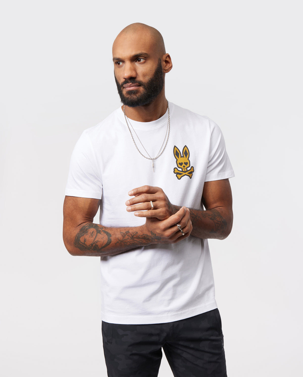 A man with a bald head and beard is wearing a white Psycho Bunny MENS VEGAS CITY TEE - B6U967X1PC in Pima cotton jersey featuring an illustrated yellow Bunny Logo and crossbones design. He has multiple tattoos on his arms and accessorizes with silver chains and rings. He stands against a plain white background.