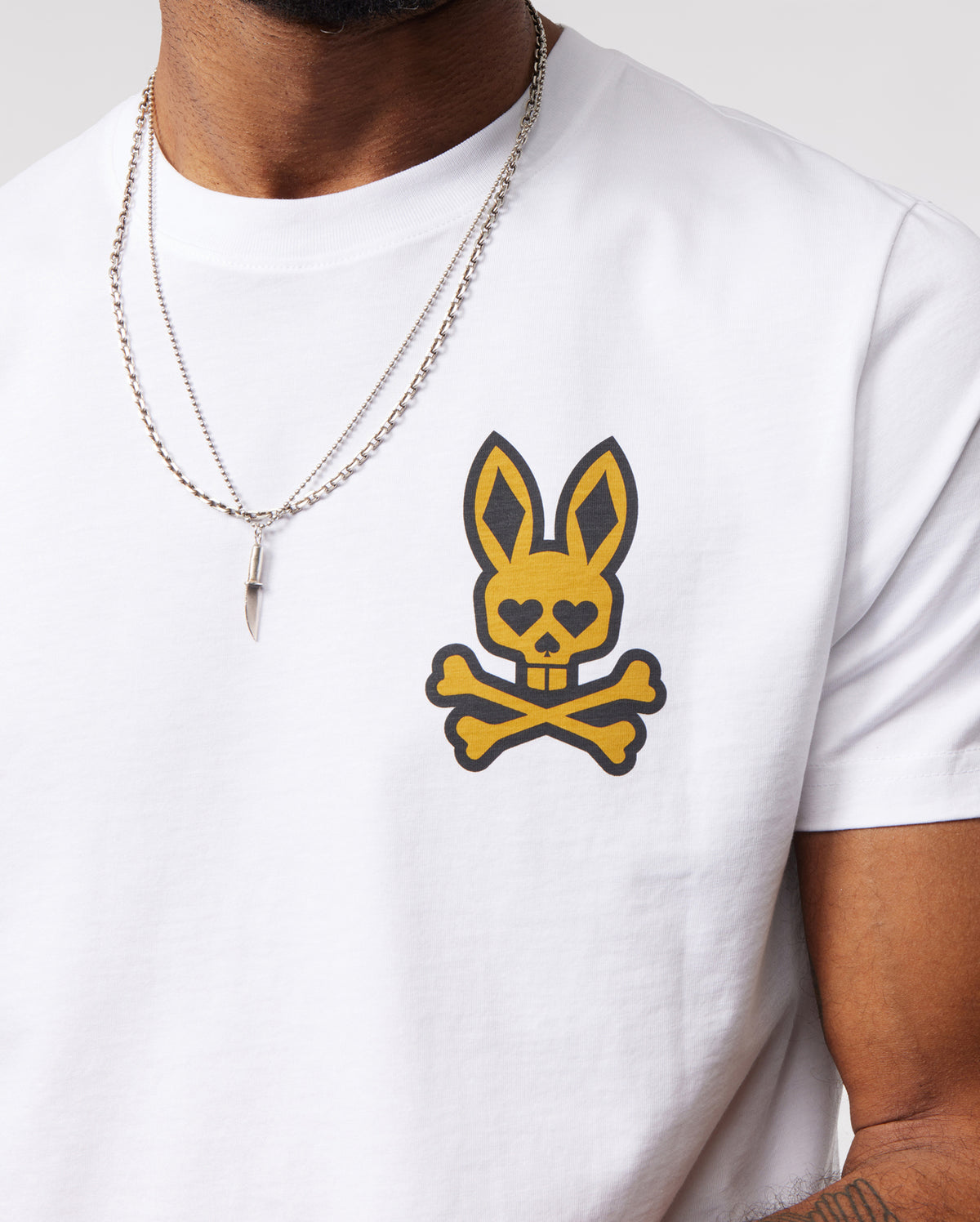 A person wearing a Psycho Bunny MENS VEGAS CITY TEE - B6U967X1PC made from soft Pima cotton jersey, featuring a yellow and black printed bunny logo on the chest. The person is also wearing a silver chain necklace with a pointed pendant. Only the torso and part of one arm are visible against a plain background.