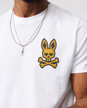 A person wearing a Psycho Bunny MENS VEGAS CITY TEE - B6U967X1PC made from soft Pima cotton jersey, featuring a yellow and black printed bunny logo on the chest. The person is also wearing a silver chain necklace with a pointed pendant. Only the torso and part of one arm are visible against a plain background.